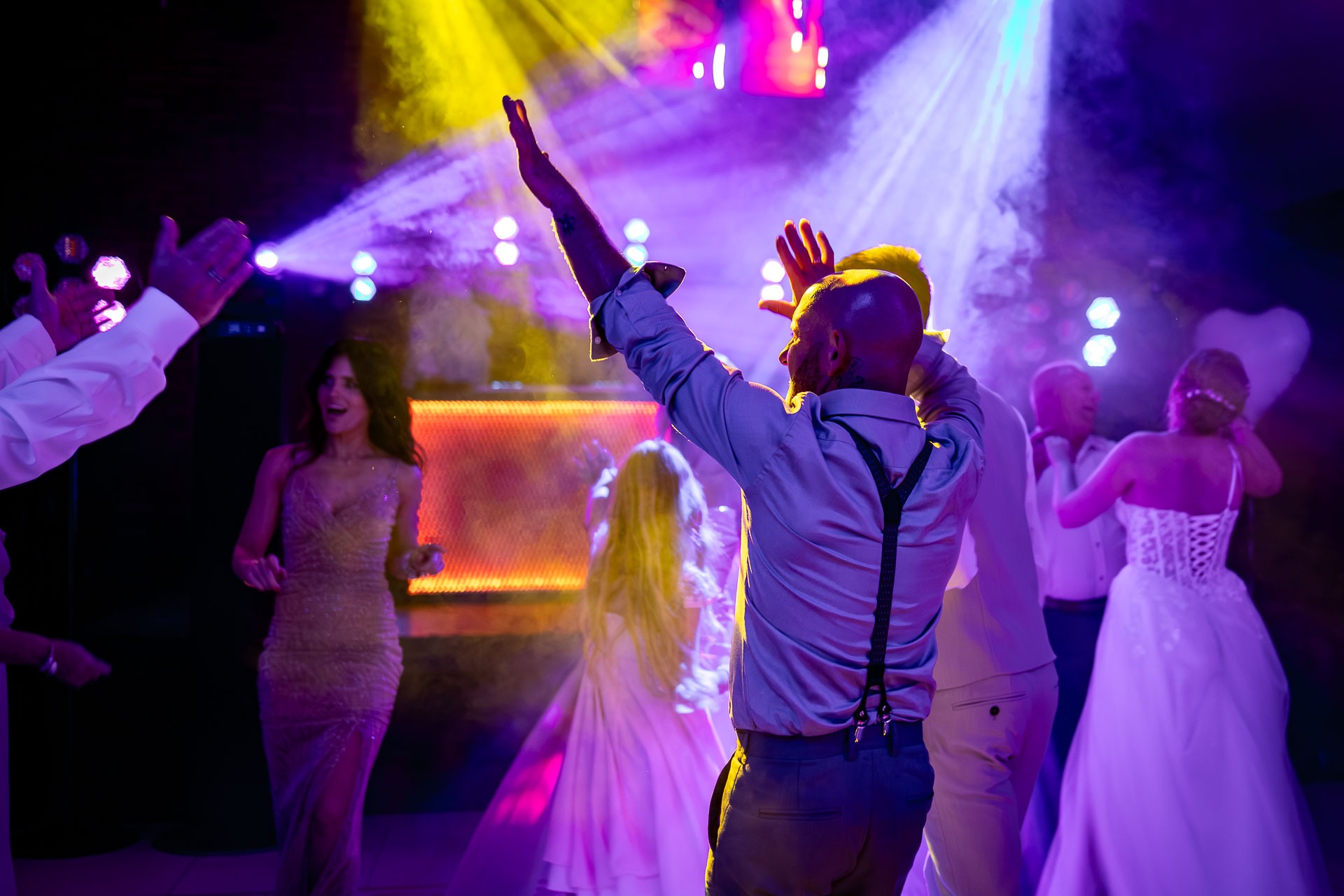 Roztańczona para młoda i goście weselni w blasku kolorowych świateł. Mężczyzna w szelkach z podniesionymi rękami. Radosna atmosfera zabawy.
