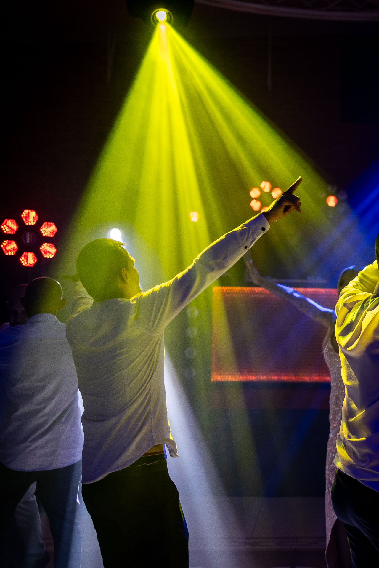 Rozświetlona scena z tańczącymi osobami w białych koszulach, pod żółtym reflektorem. Dynamiczne ujęcie z podniesioną ręką i widocznymi światłami scenicznymi w tle.