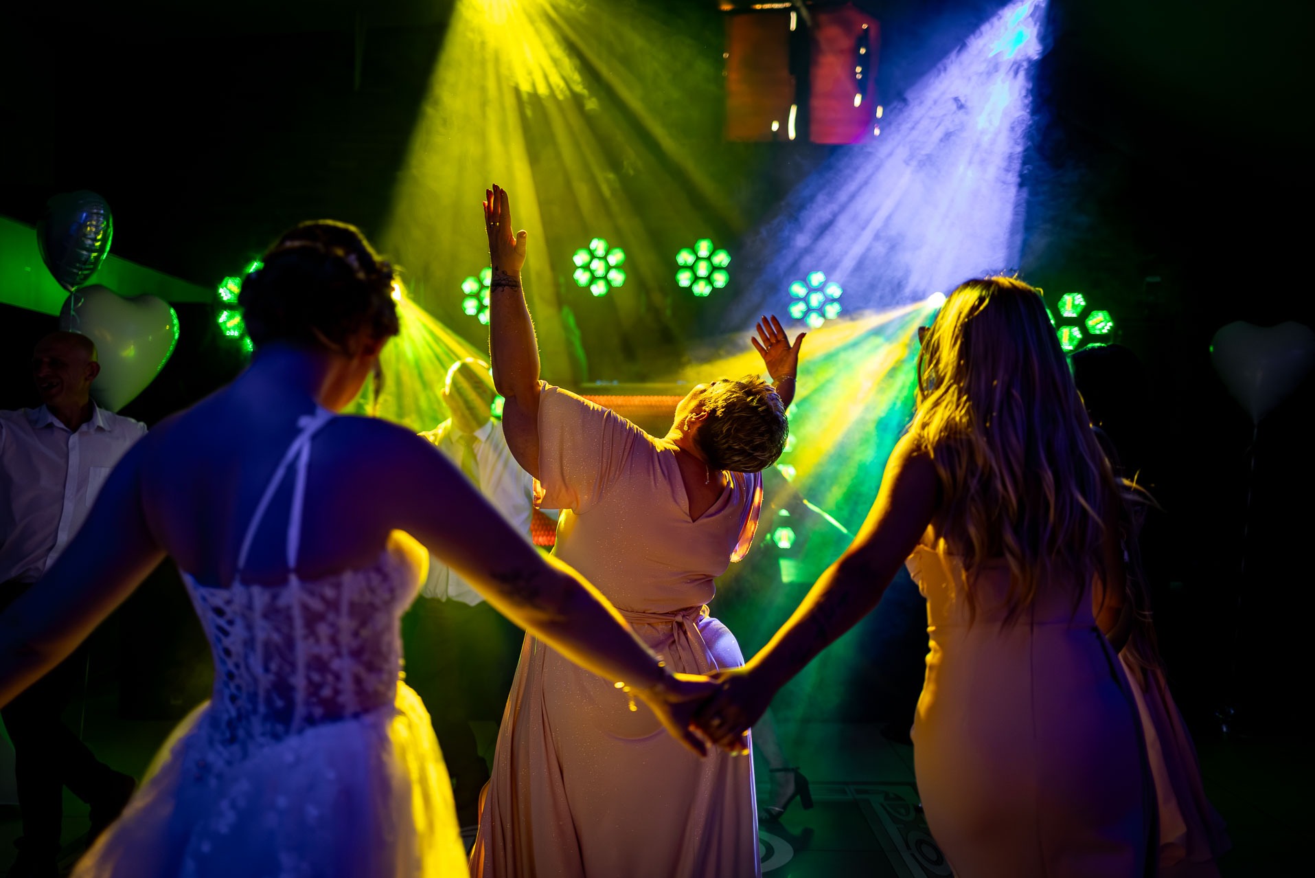 Rozświetlona sala balowa, goście weselni w tańcu, widok zza pleców panny młodej w koronkowej sukni, dynamiczne oświetlenie sceniczne i dekoracje.