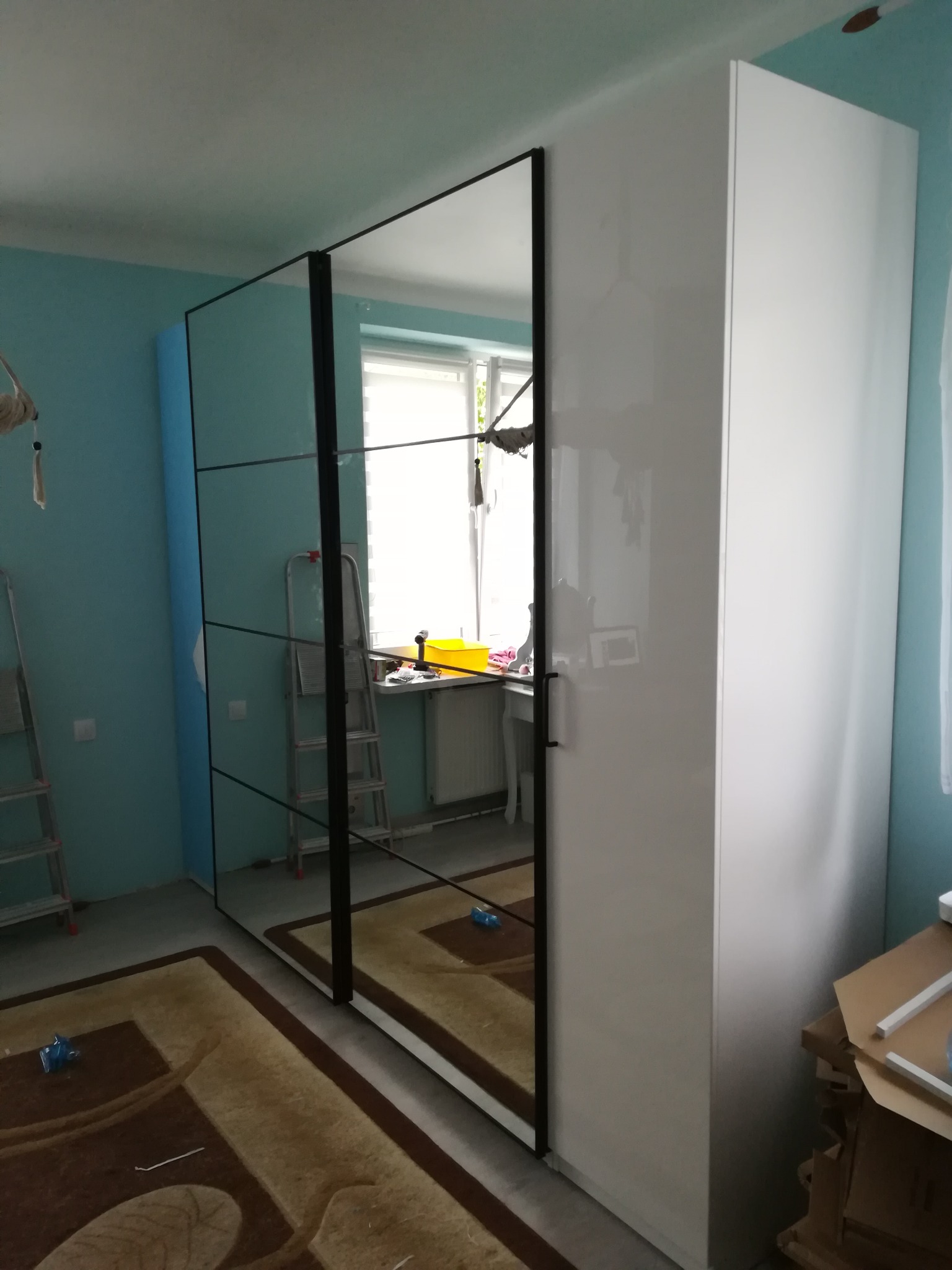 Wysoka, biała szafa PAX z przesuwanymi drzwiami z lustrami w czarnych ramach, widoczna w pokoju z turkusowymi ścianami i dywanem w odcieniach brązu, z elementami montażu widocznymi w tle.