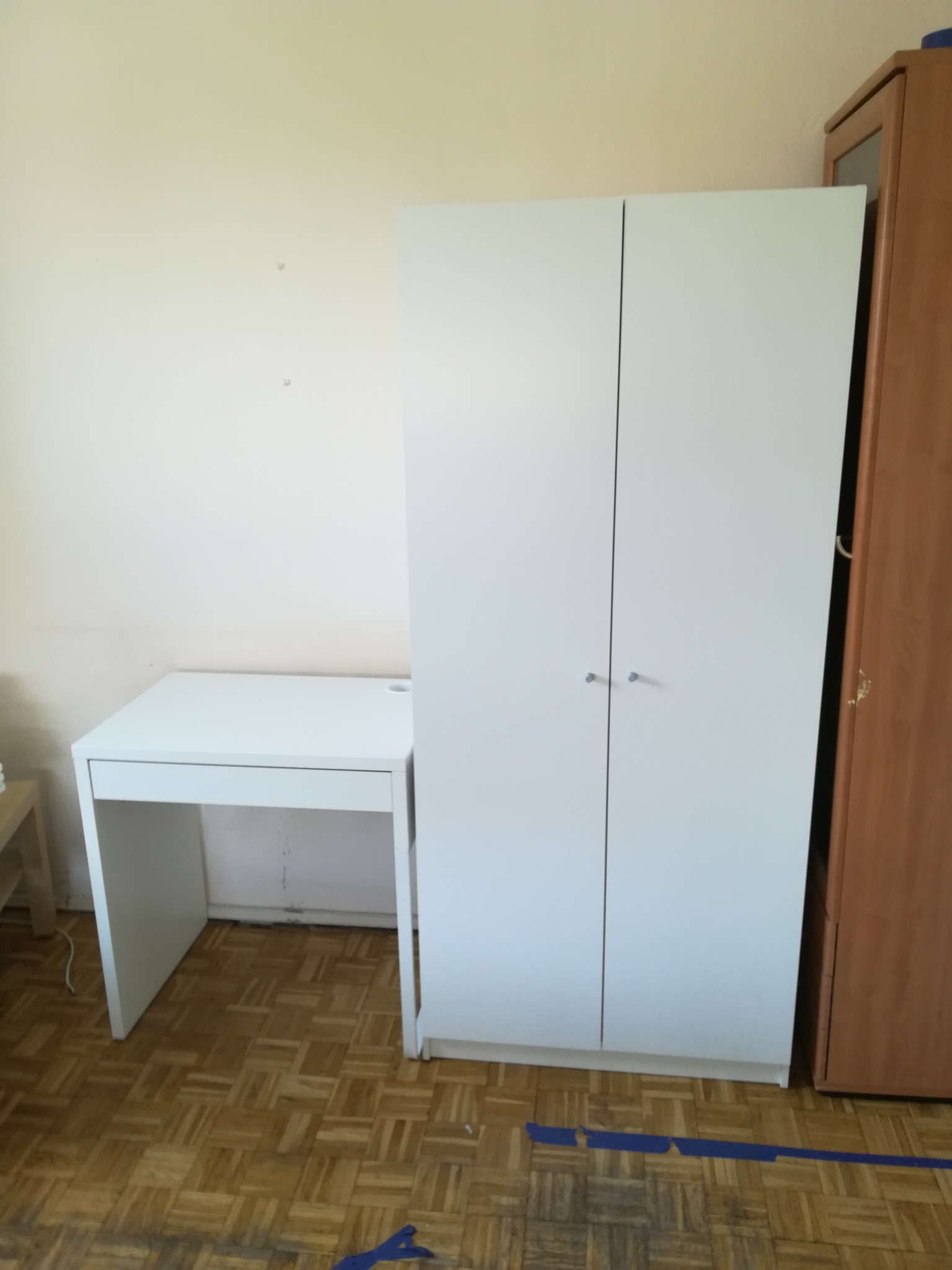 Białe biurko i szafa z zamontowanymi drzwiami w pokoju z parkietem, widoczna taśma malarska na podłodze.
