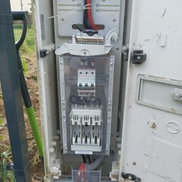 MWAInstal - Otwarta skrzynka elektryczna z widocznymi bezpiecznikami, przewodami i elementami montażowymi. Widoczne ślady użytkowania i montażu w terenie.