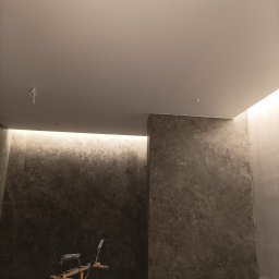 MWAInstal - Podświetlona ściana z marmuru w łazience, widoczny reflektor budowlany, sufit z punktowym oświetleniem. Wykończenie wnętrza w trakcie prac.