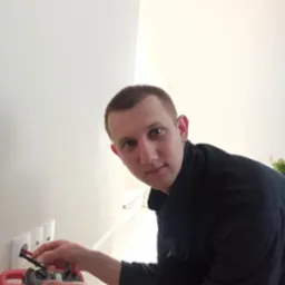 Elektryk mierzy napięcie w gniazdku elektrycznym za pomocą miernika cęgowego, na tle białej ściany.