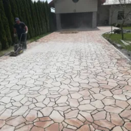 Układanie nawierzchni z nieregularnych, jasnych kamieni z ciemną fugą. Pracownik ubija nawierzchnię zagęszczarką w pobliżu garażu i żywopłotu.