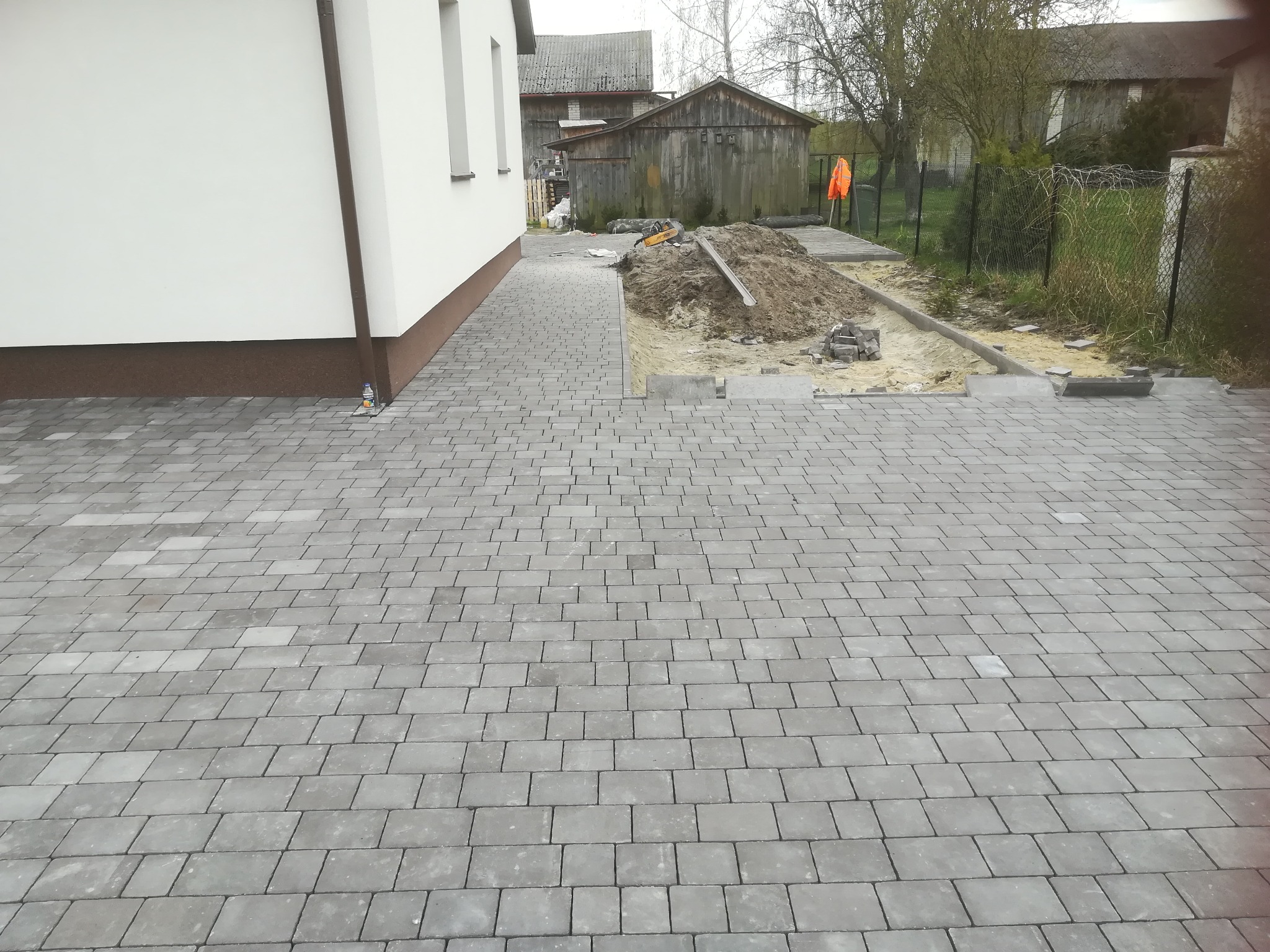 Ułożona szara kostka brukowa na podjeździe, widoczny fragment budynku z białymi ścianami i brązowym cokołem, w tle sterta ziemi i przygotowane obrzeża pod dalszą część nawierzchni.