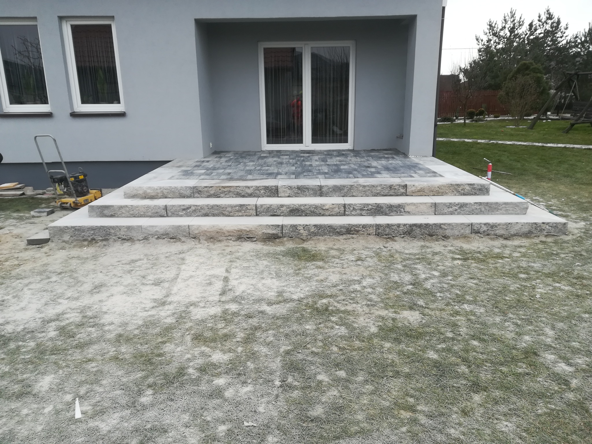 Wykończone schody wejściowe z kostki brukowej prowadzące do domu z szarymi ścianami, widoczna zagęszczarka w tle.