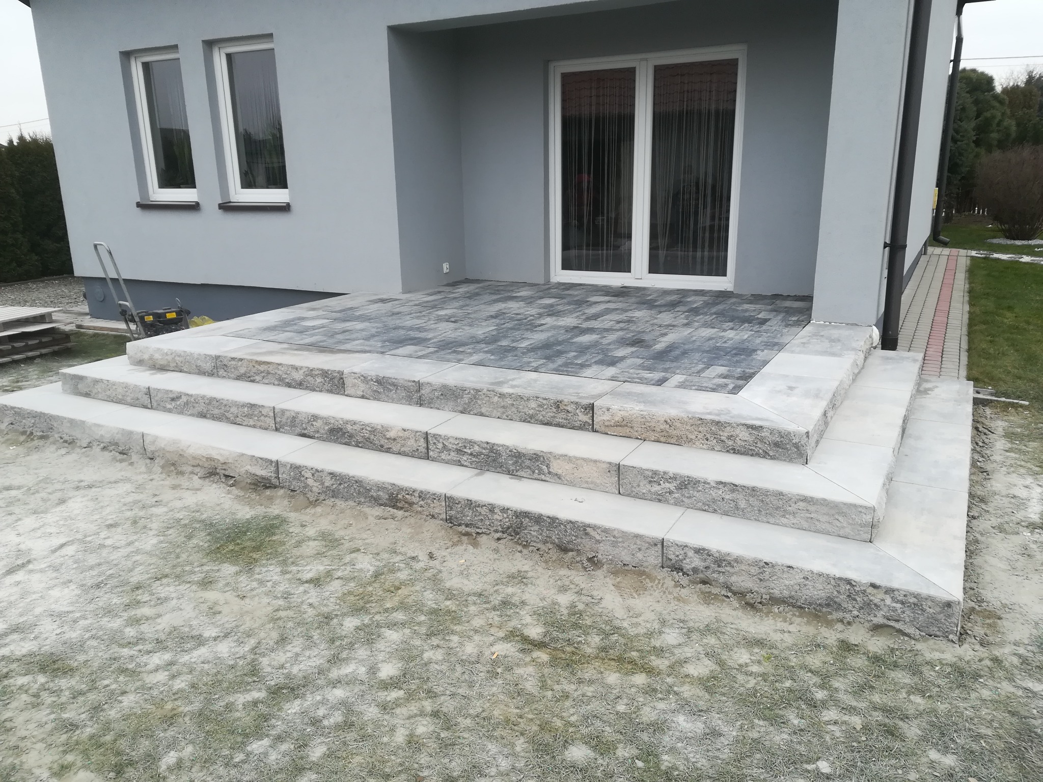 Schody wejściowe do szarego domu wyłożone jasnoszarą kostką, z podjazdem z kostki w odcieniach szarości i czerni. W tle widoczne okna i drzwi balkonowe.