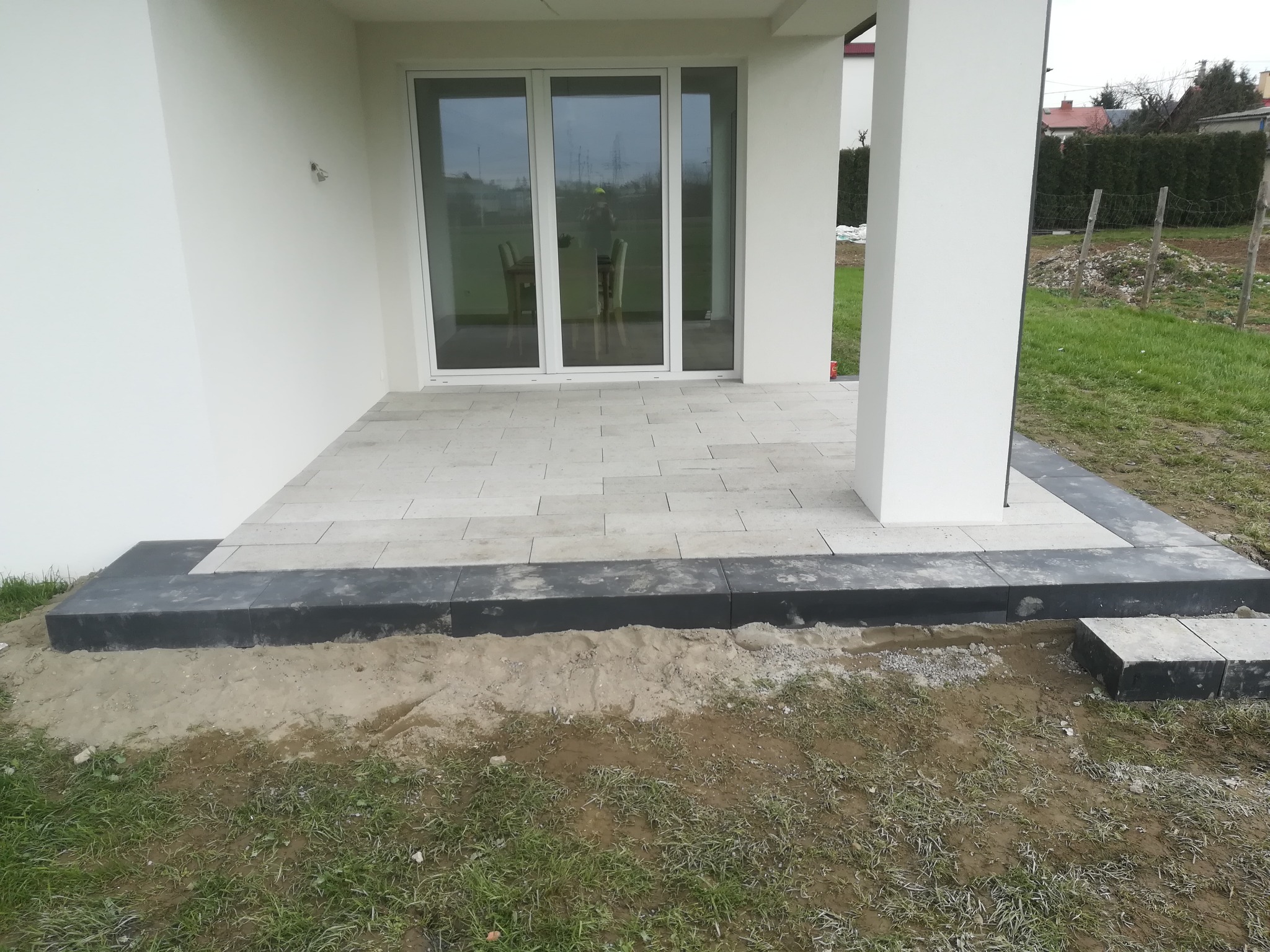 Jasnoszara kostka ułożona przed wejściem do domu z białymi ścianami i dużymi przesuwnymi drzwiami, obrzeże z ciemnej kostki, widoczny fragment trawnika i ziemi.