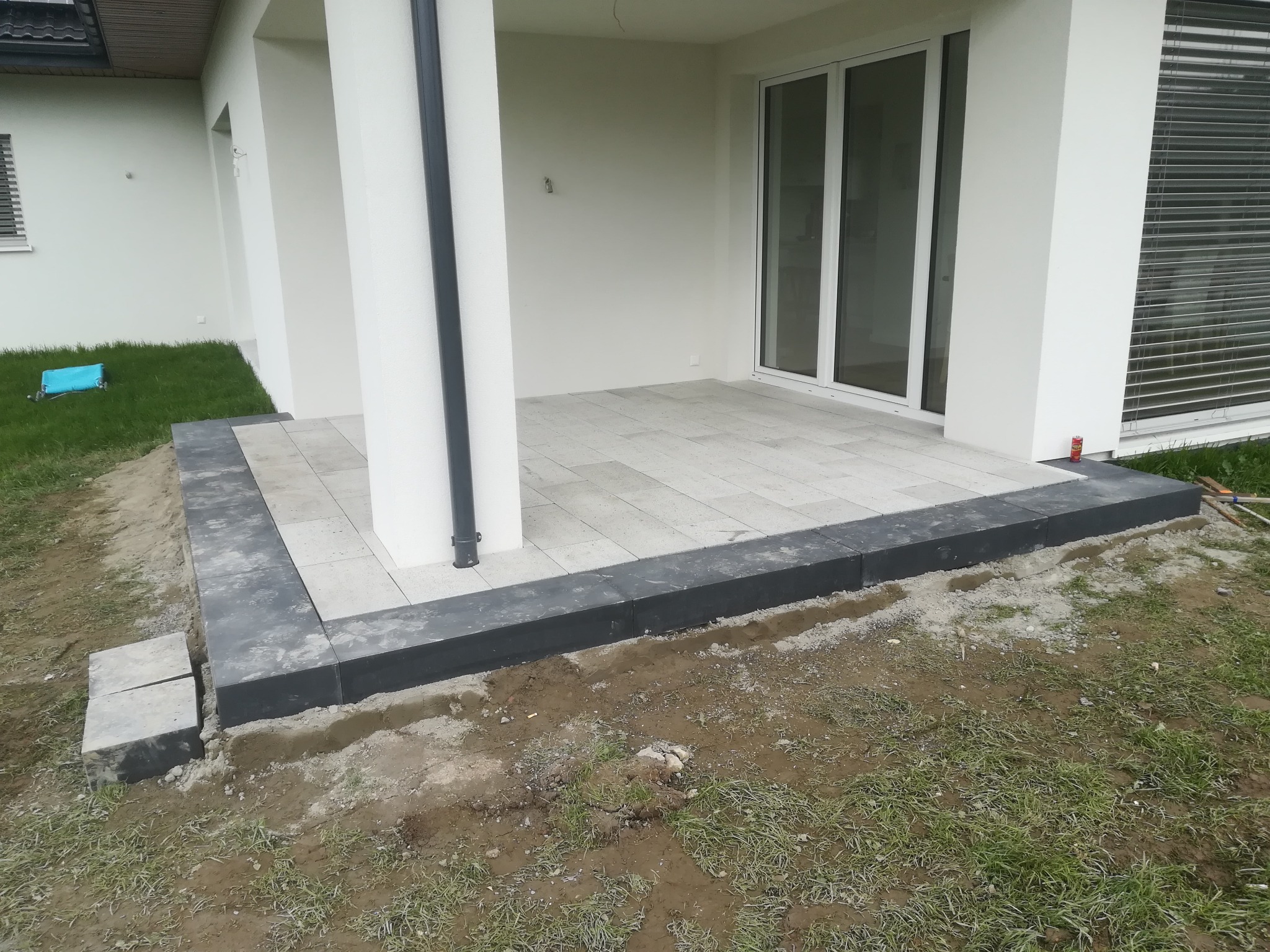 Wyłożony jasnoszarą kostką brukową taras przy nowoczesnym domu z czarną obwódką, widoczne niedokończone prace ziemne przy krawędziach.