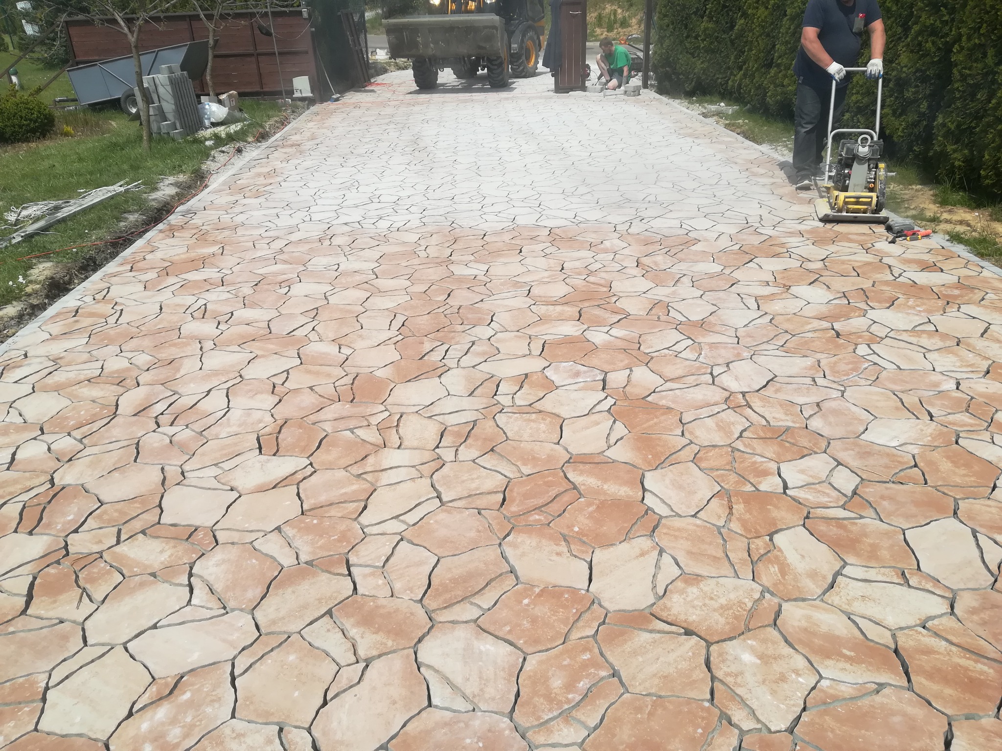 Układanie nieregularnej kostki w odcieniach beżu i brązu, zagęszczarka wibrująca w użyciu, widoczny operator i sprzęt budowlany w tle.