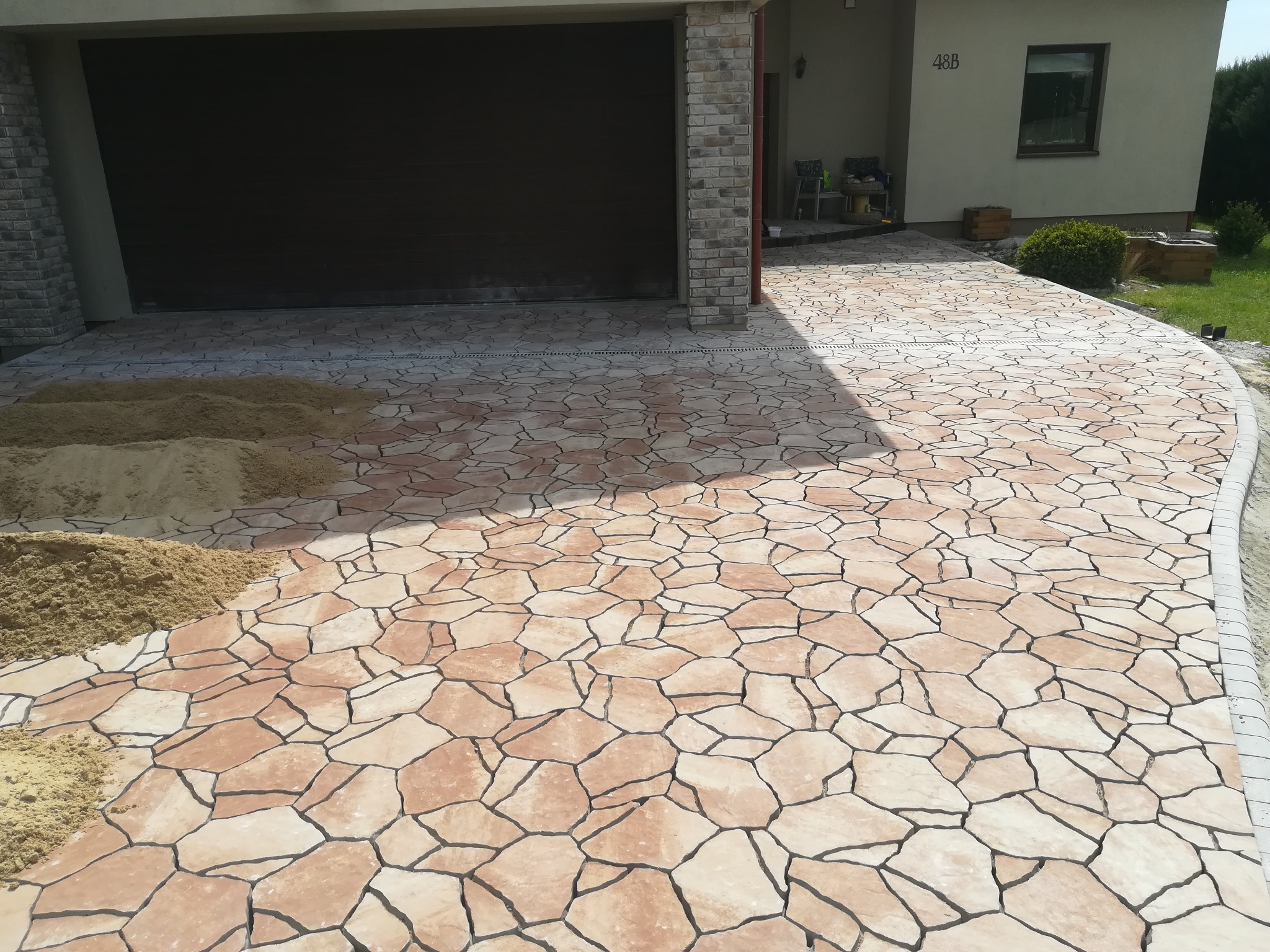 Nowa nawierzchnia z nieregularnych, beżowo-brązowych płyt kamiennych prowadząca do garażu i domu, z widocznymi hałdami piasku przygotowanymi do zasypania szczelin.