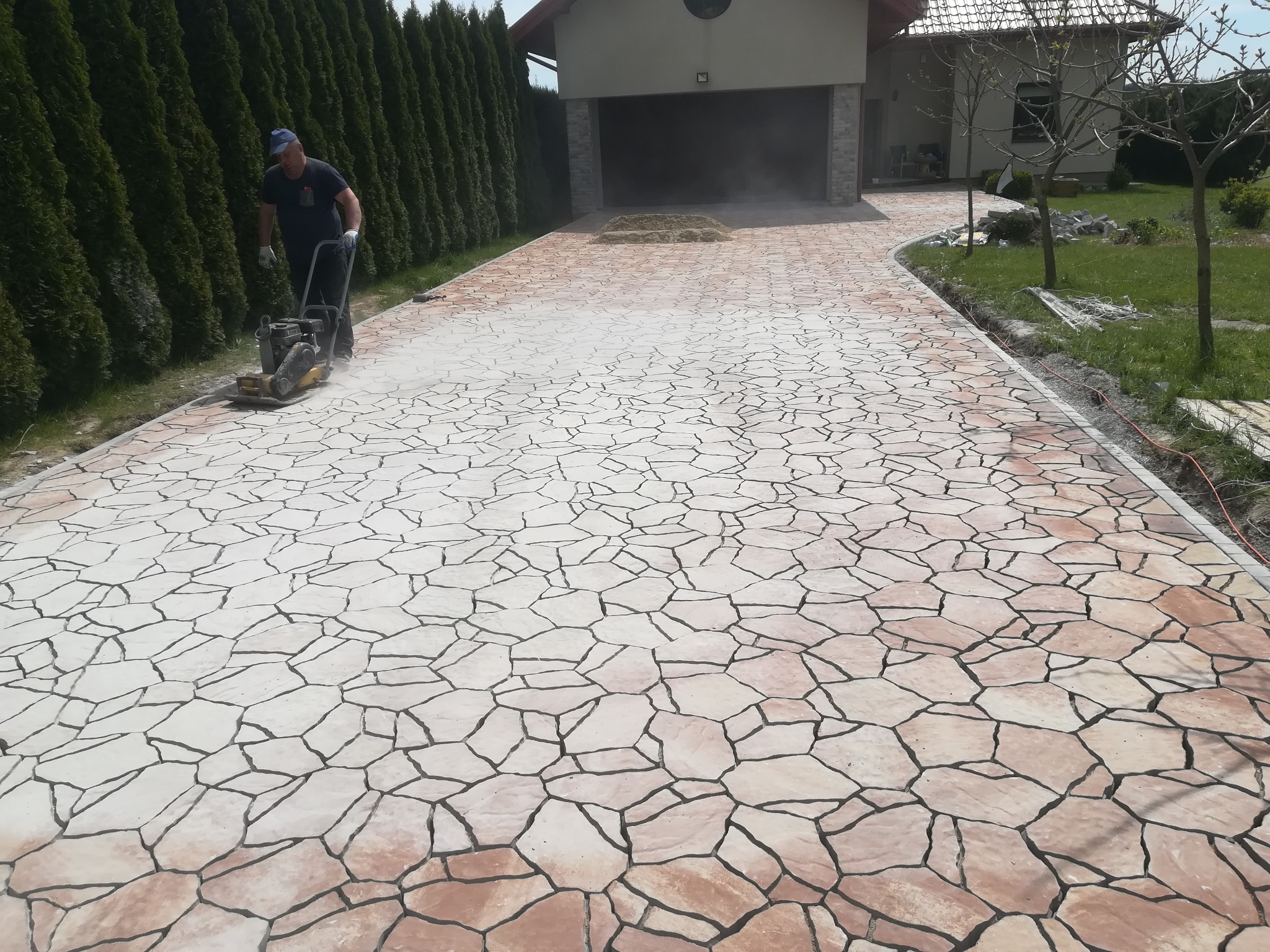 Układanie nawierzchni z nieregularnych, jasnych kamieni z ciemną fugą. Pracownik ubija nawierzchnię zagęszczarką w pobliżu garażu i żywopłotu.