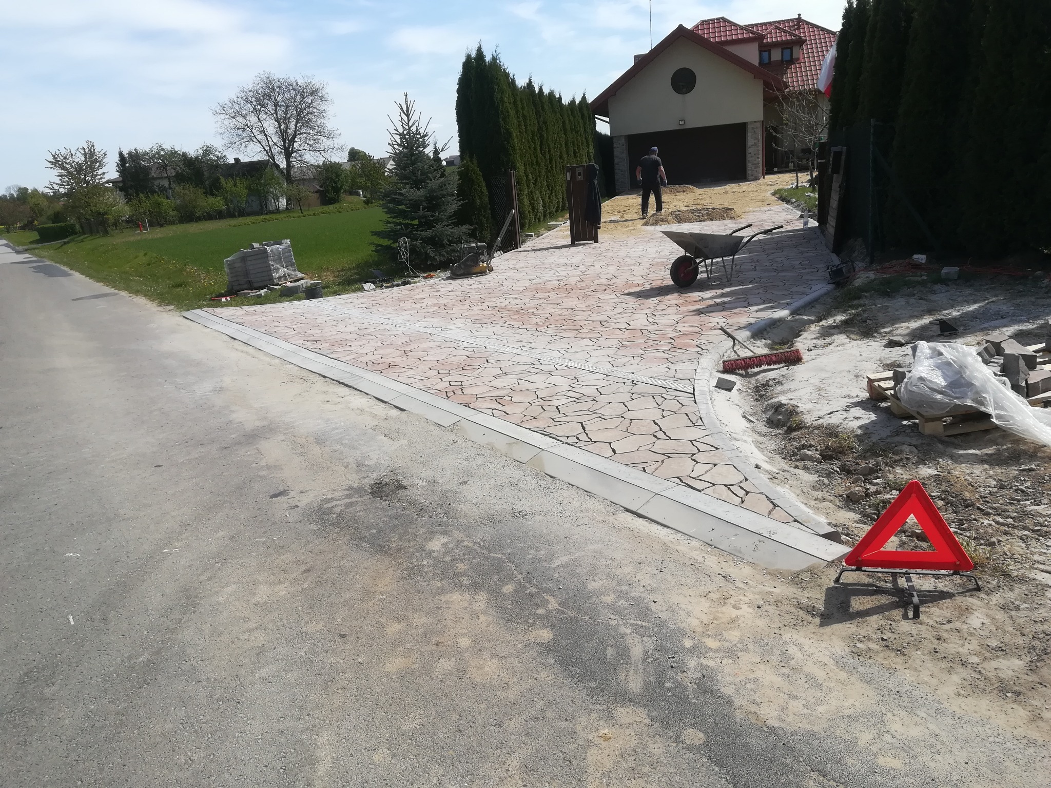 Ułożona ścieżka z nieregularnej kostki na posesji, widoczny pracownik z taczką, narzędzia i trójkąt ostrzegawczy na poboczu.