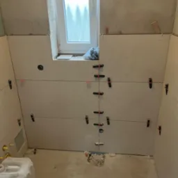 Łazienka w trakcie układania beżowych płytek ściennych, widoczne czarne klipsy poziomujące, okno z widokiem na zewnątrz, surowy beton na podłodze.