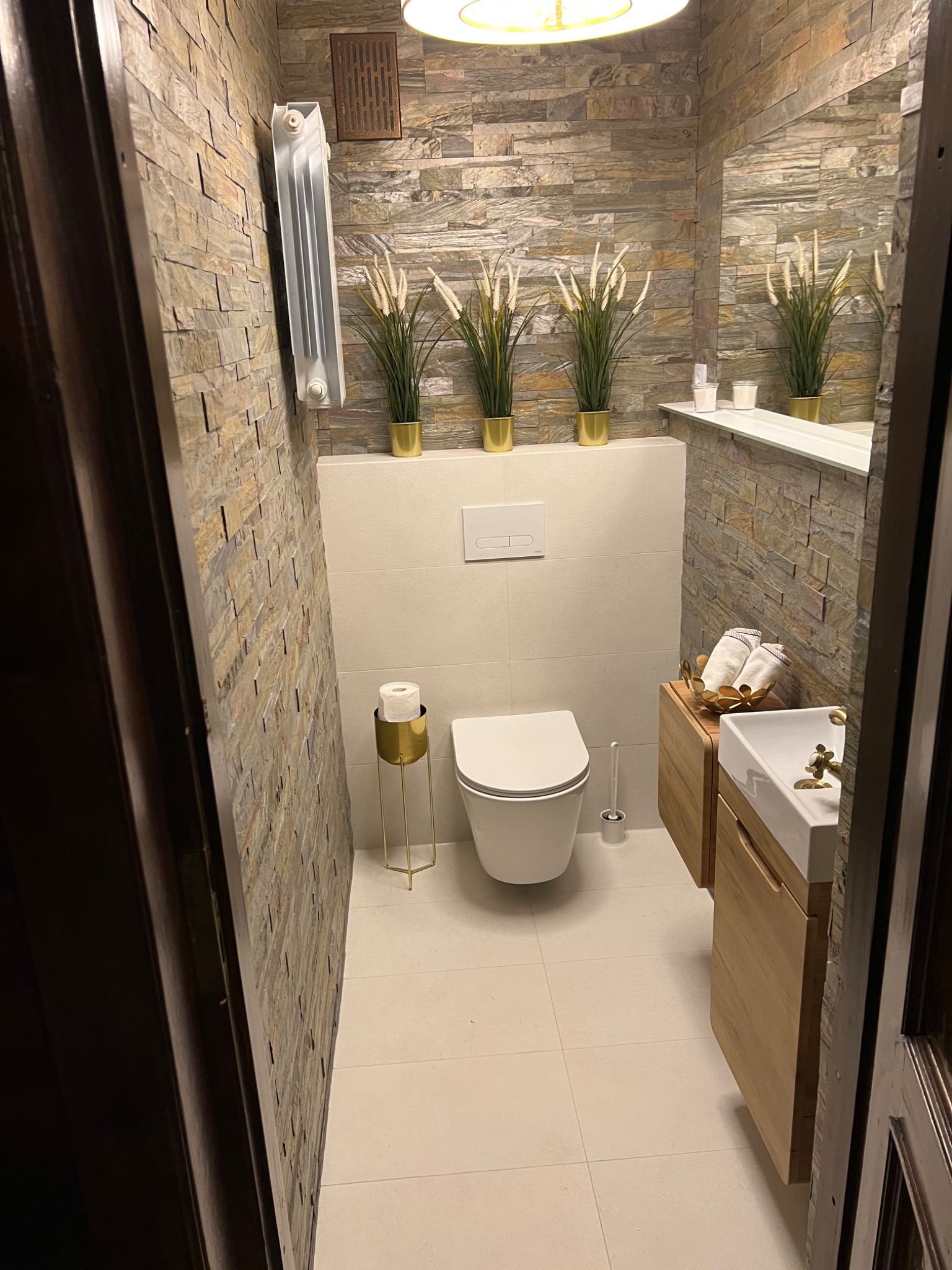 Nowoczesna toaleta z kamienną ścianą, wiszącą muszlą klozetową, umywalką nablatową na drewnianej szafce, złotymi akcentami i dekoracyjnymi trawami w złotych doniczkach.
