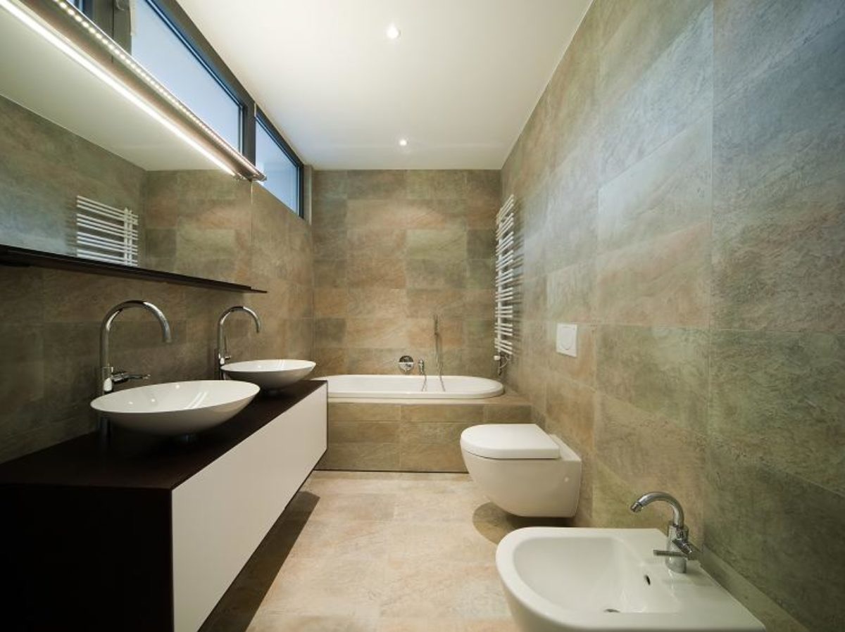 Nowoczesna łazienka z dwoma umywalkami na blacie, wanną w zabudowie, wiszącą toaletą i bidetem, wykończona płytkami w odcieniach beżu i szarości, oświetlona listwą LED.