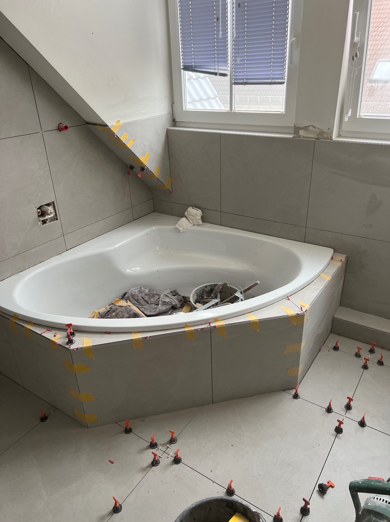 Narożna wanna obłożona szarymi płytkami, z widocznymi elementami poziomującymi i taśmą maskującą, odzwierciedlająca etap prac wykończeniowych w łazience.