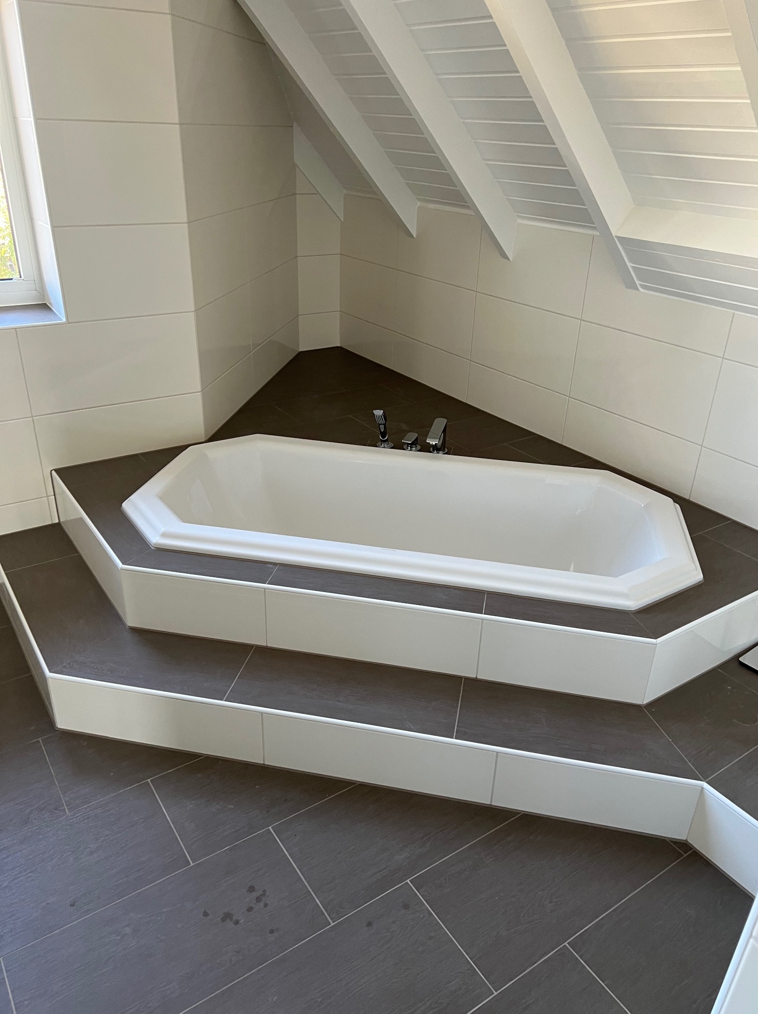 Wanna w zabudowie na podwyższeniu, wykończona ciemnymi płytkami imitującymi drewno, białe płytki na ścianach i skośnym suficie z drewnianą boazerią.