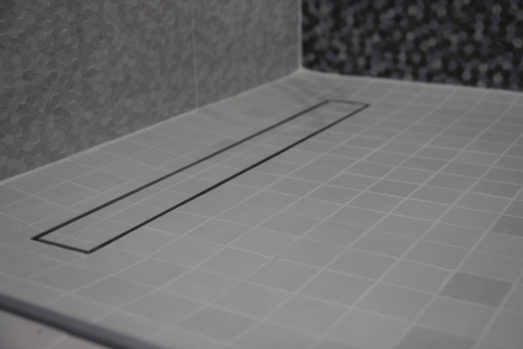 Białe płytki podłogowe w łazience z czarnym odpływem liniowym, fragment ściany wykończony mozaiką.