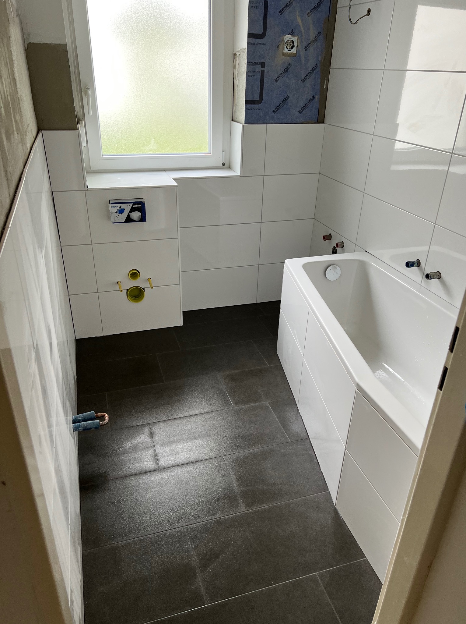 Łazienka w trakcie remontu: położone białe płytki ścienne, ciemnoszara podłoga, wanna obudowana płytkami, widoczne przygotowane przyłącza wodne i kanalizacyjne, częściowo odkryta ściana z izolacją.