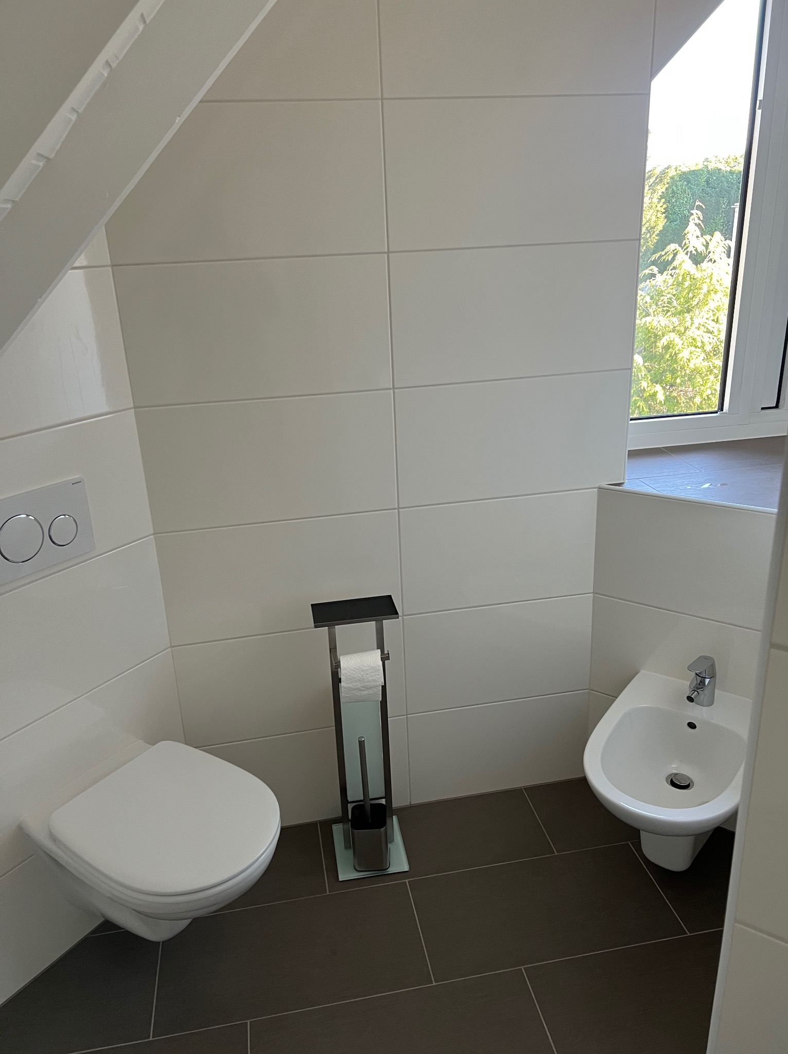 Nowoczesna łazienka na poddaszu z białymi płytkami, wiszącą toaletą, bidetem i ciemną podłogą z płytek. Widok na zieleń za oknem.