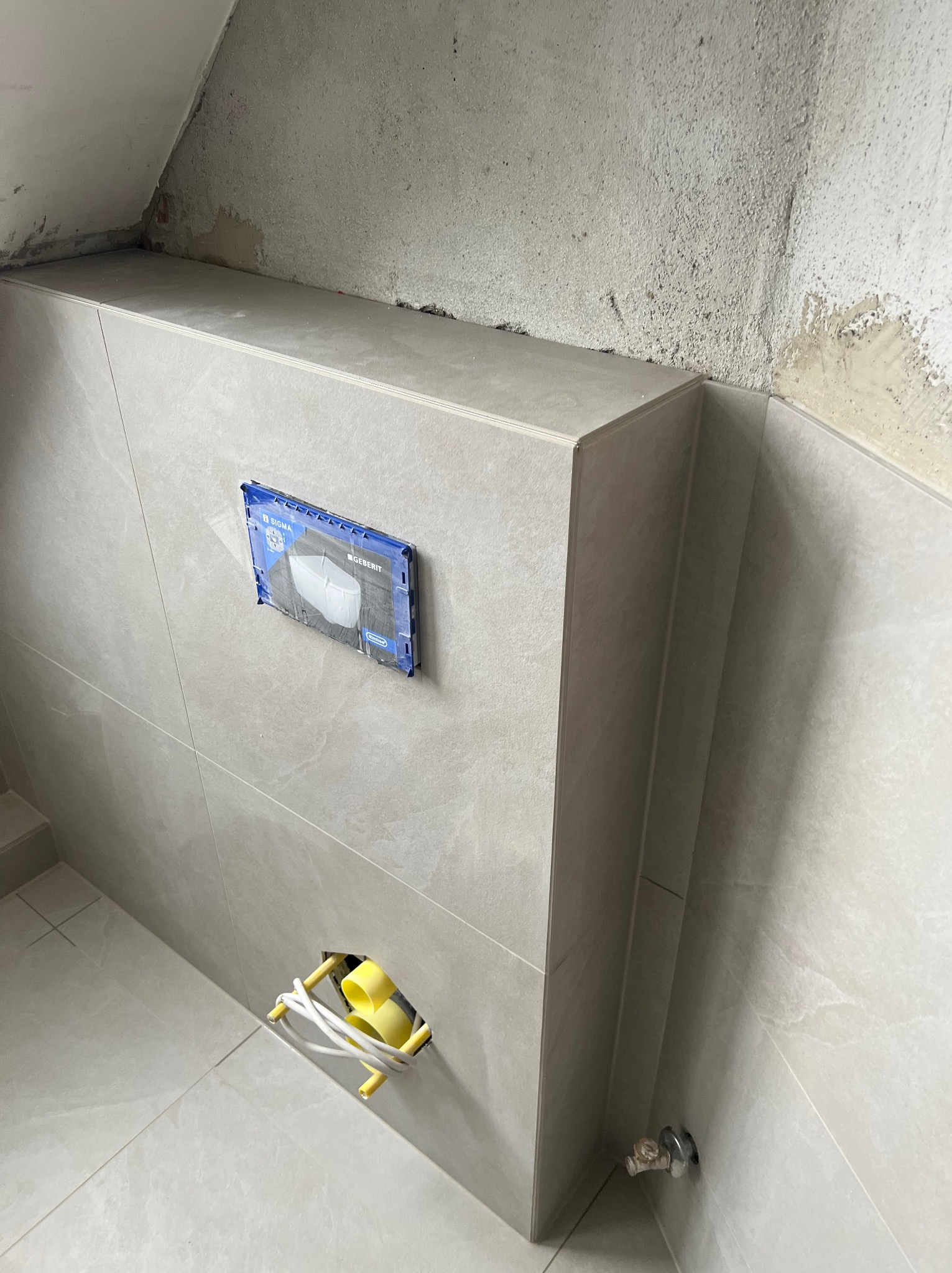 Ściana łazienki w trakcie prac wykończeniowych, widoczne szare płytki, zabudowa stelaża podtynkowego oraz wystające przewody elektryczne i instalacja wodna.