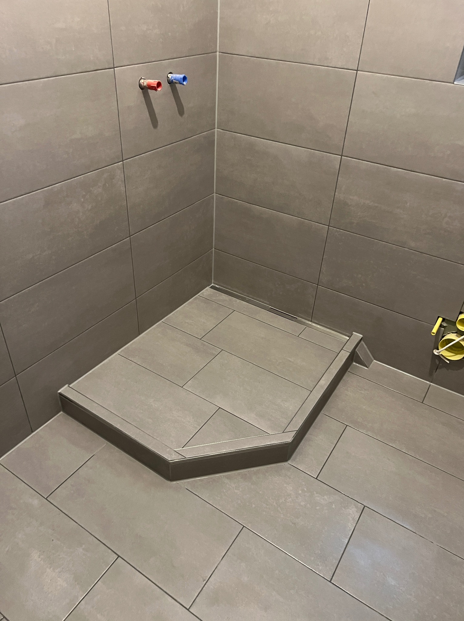 Kafelkowany brodzik bez kabiny z odpływem liniowym i wystającymi rurami ciepłej i zimnej wody w łazience wykończonej płytkami w odcieniach szarości.