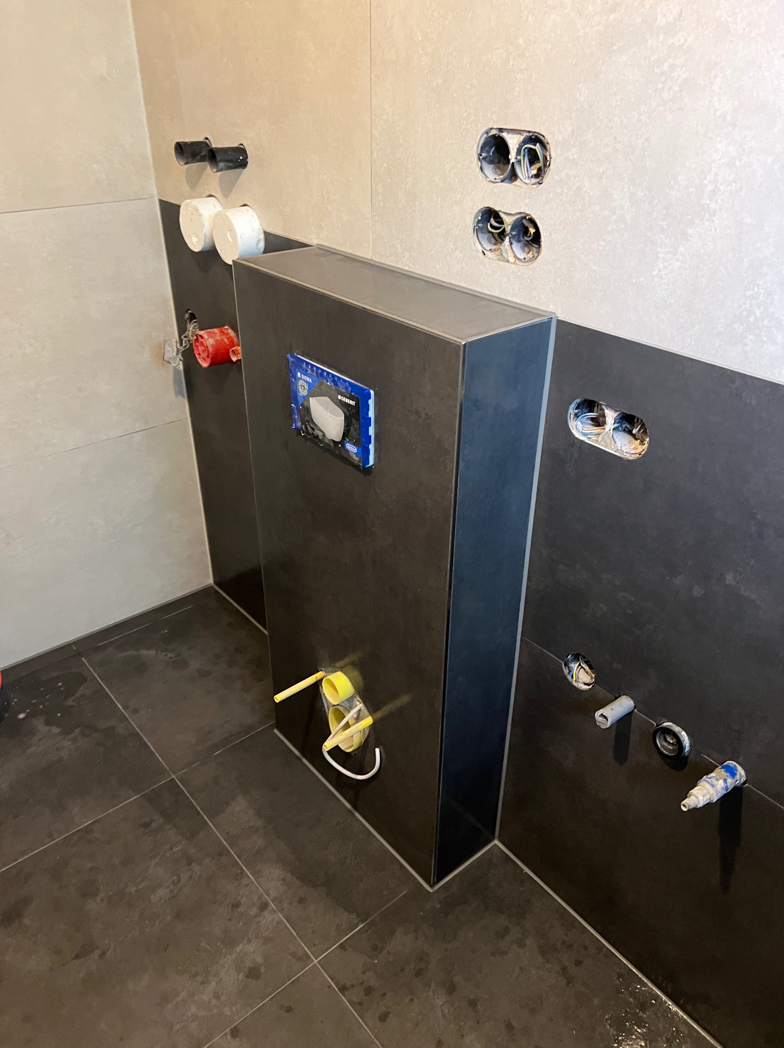 Montaż stelaża podtynkowego WC z widocznymi przyłączami wodno-kanalizacyjnymi i elektrycznymi w łazience wykończonej płytkami w odcieniach szarości.