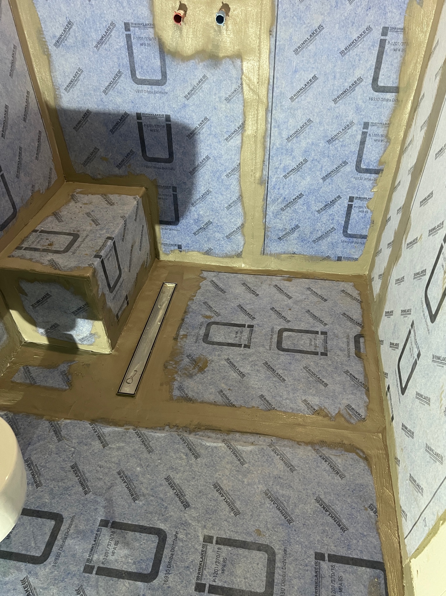 Przygotowanie do hydroizolacji w łazience: ściany i podłoga pokryte niebieską membraną w płynie z widocznym odpływem liniowym i fragmentem toalety.