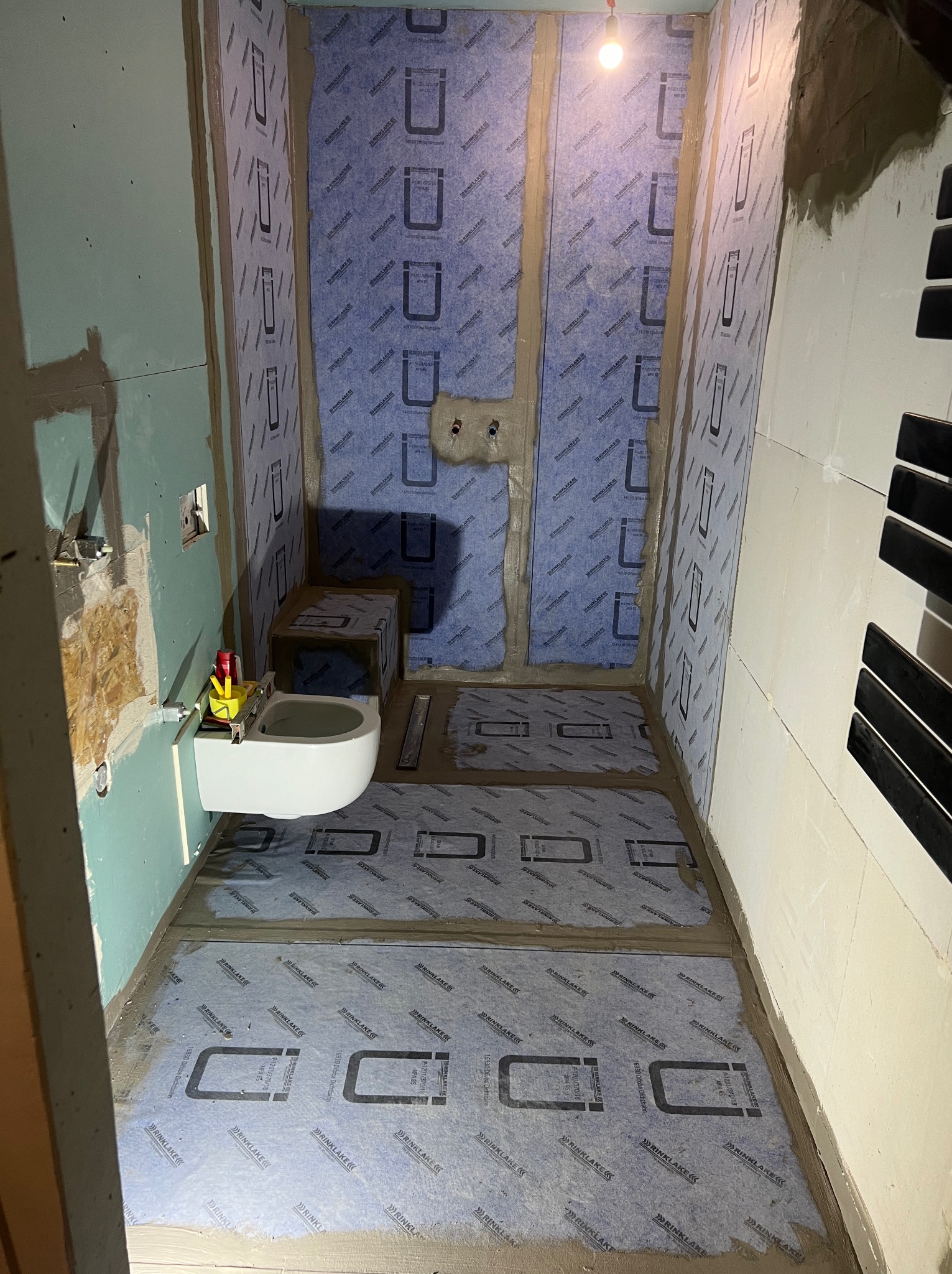 Łazienka w trakcie prac: hydroizolacja podłoża z zamontowaną miską WC podwieszaną, widoczne narzędzia i materiały budowlane.
