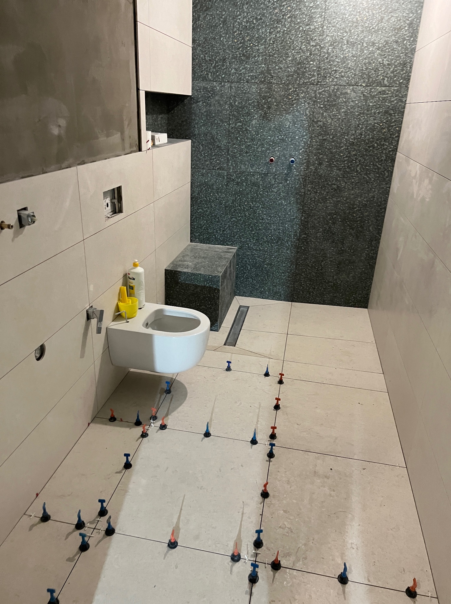 Łazienka w trakcie wykańczania: białe płytki na ścianach, ciemne mozaiki na jednej ścianie, biała wisząca miska WC z żółtym wiaderkiem i butelką płynu, poziomowanie płytek podłogowych za pomocą...