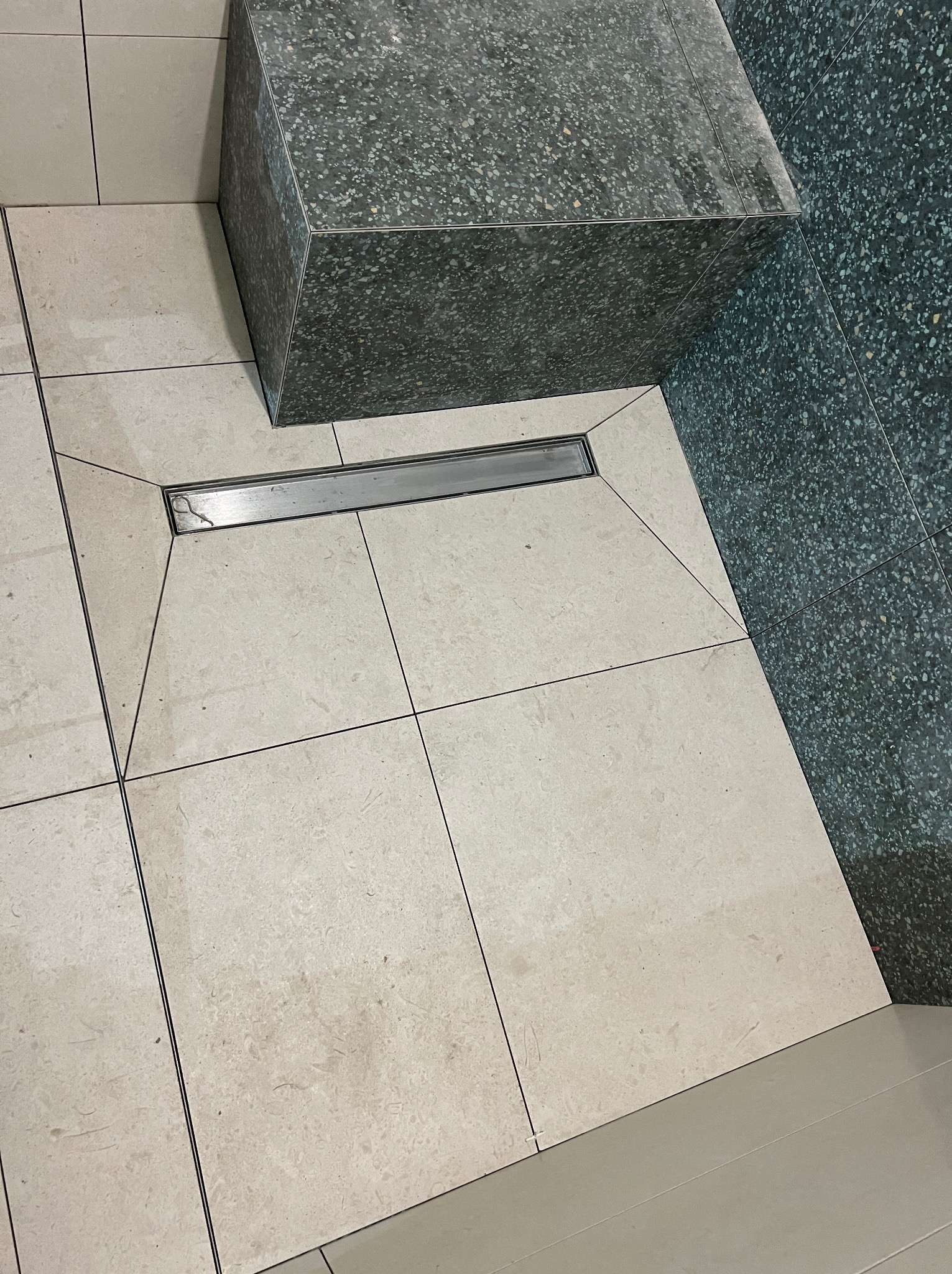 Beżowe płytki podłogowe w łazience z odpływem liniowym i fragmentem obudowy z ciemnego lastryko