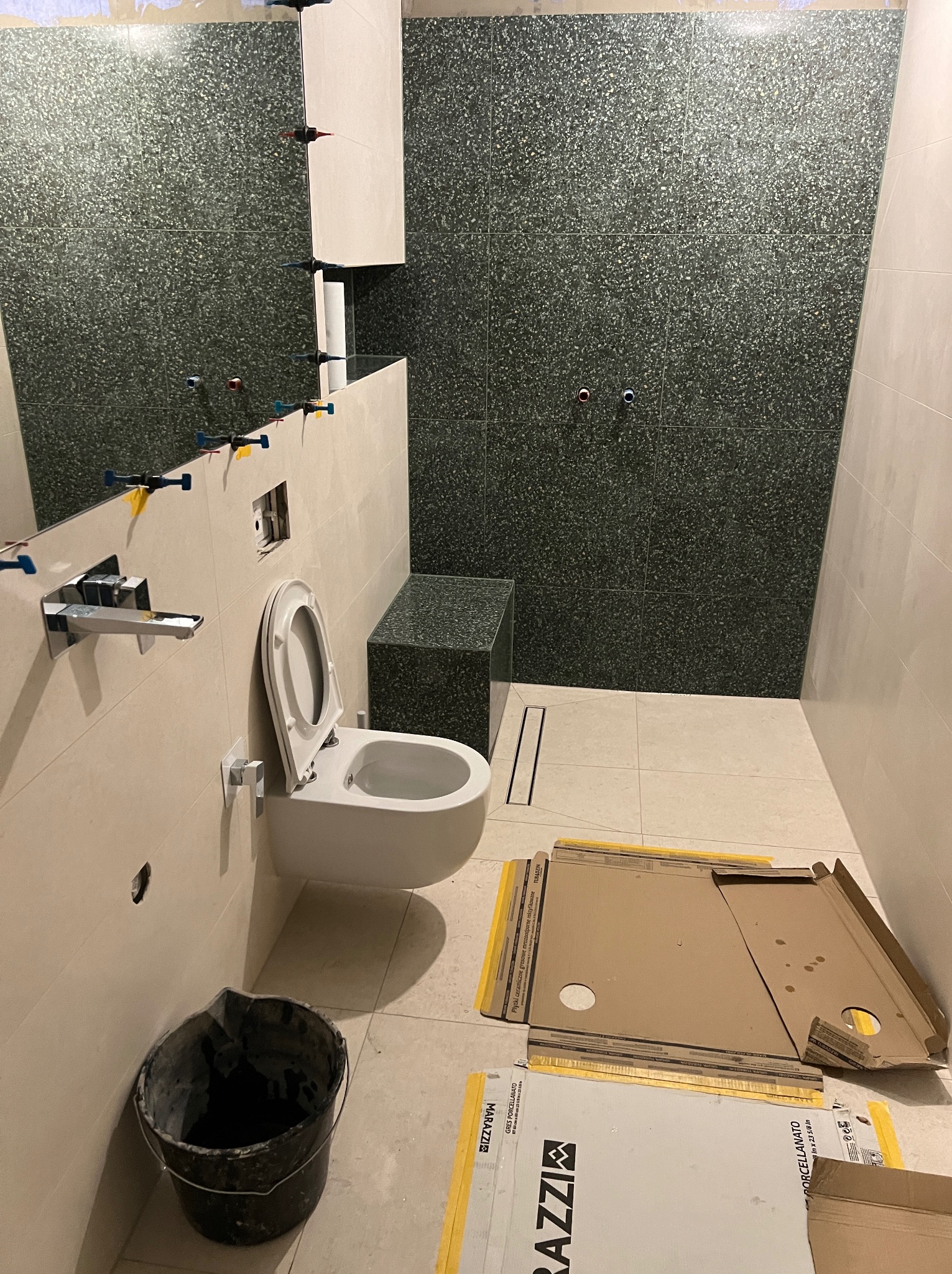 Remont łazienki w trakcie: położone płytki w odcieniach szarości na ścianach, biała wisząca toaleta z otwartą klapą, widoczne narzędzia glazurnicze i kartony po płytkach Marazzi na podłodze.