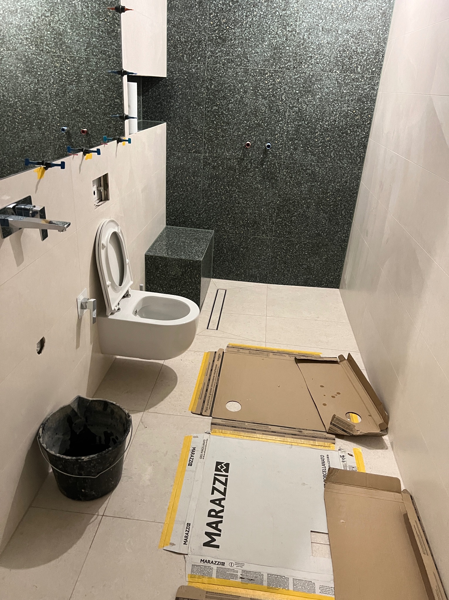 Łazienka w trakcie wykańczania: podwieszana toaleta, częściowo ułożone płytki na ścianie z użyciem krzyżyków dystansowych, ciemna ściana z mozaiki, kartony po płytkach Marazzi na podłodze, wiadro...