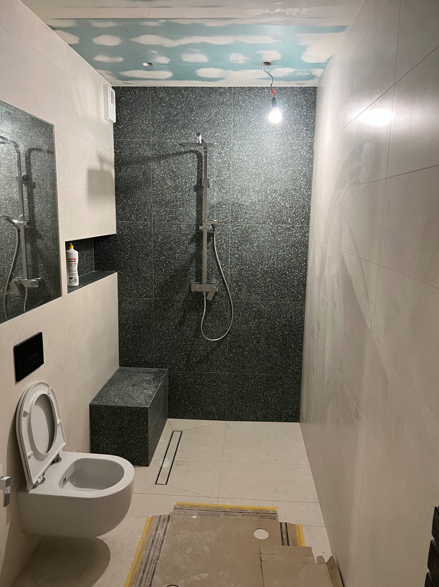 Niedokończona łazienka z ciemną mozaiką pod prysznicem, jasnymi płytkami na ścianach, podwieszaną toaletą i sufitem z efektem chmur, widoczne kartony na podłodze.