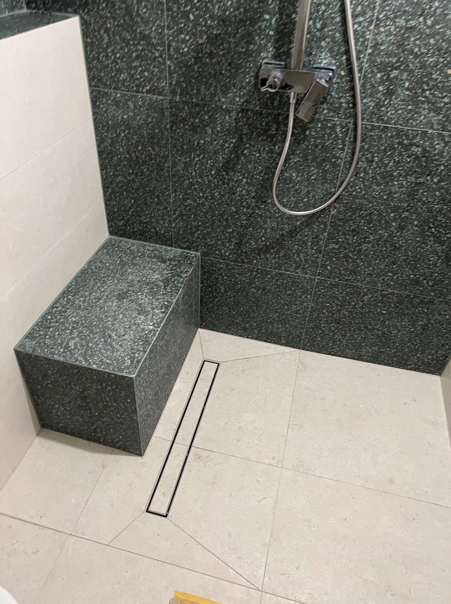 Nowoczesna kabina prysznicowa z siedziskiem wykończona płytkami w odcieniach szarości, z odpływem liniowym i chromowaną baterią.