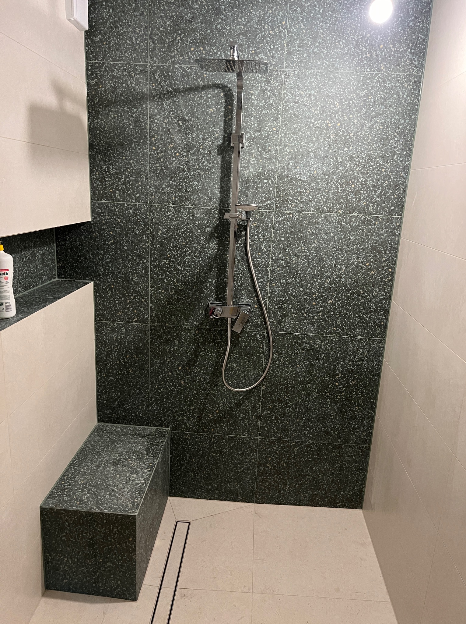 Nowoczesna kabina prysznicowa z deszczownicą, wykończona ciemnozielonymi płytkami imitującymi lastryko, z wbudowanym siedziskiem z tego samego materiału i liniowym odpływem w podłodze.