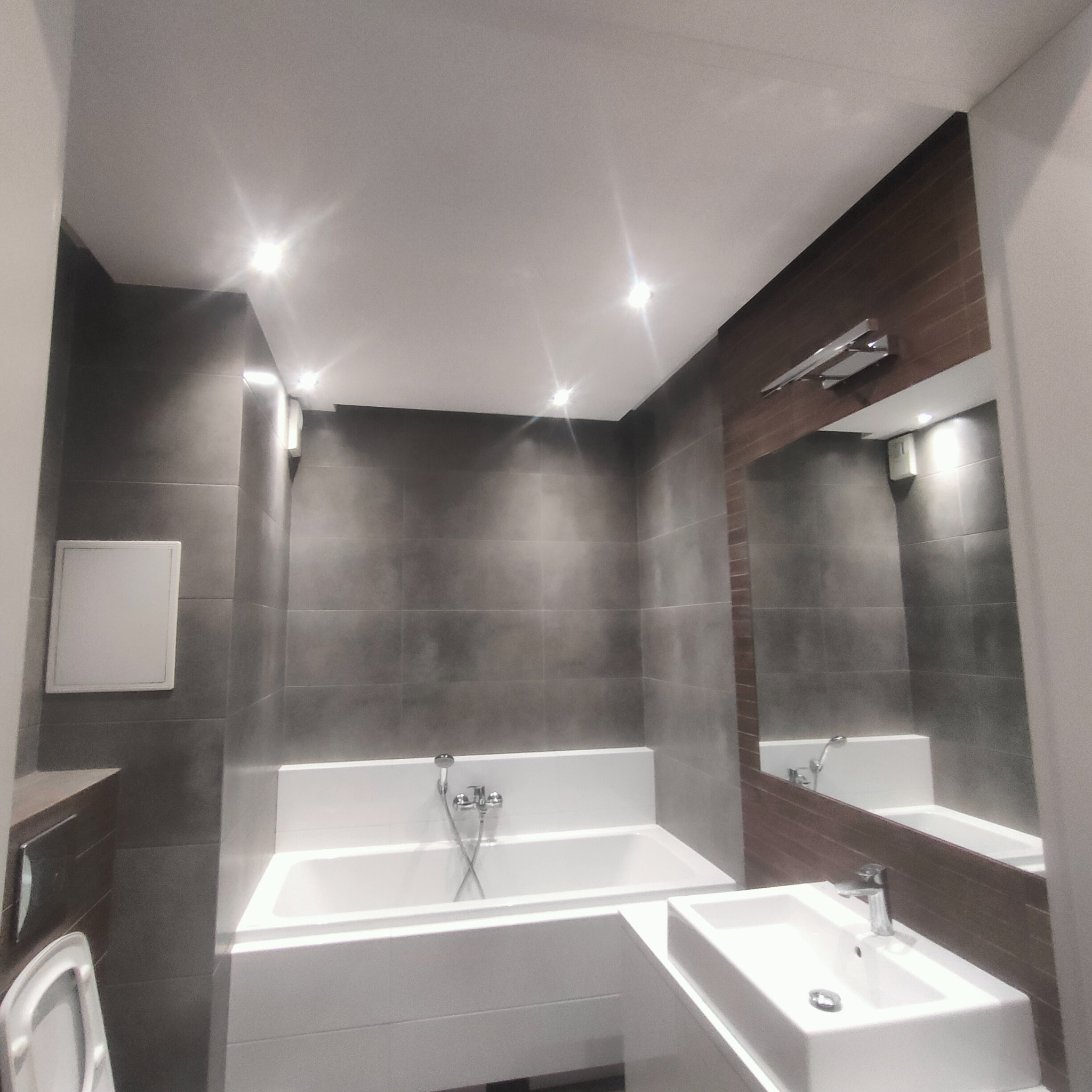 Nowoczesna łazienka z szarymi płytkami, białą wanną i umywalką, oświetleniem punktowym i dużym lustrem z drewnianym akcentem.