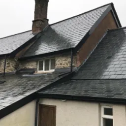 Nowo położony, mokry, ciemnoszary dach z łupka na budynku z kamienną elewacją i białymi oknami, widoczne rynny w kolorze czarnym oraz komin z cegły.