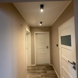 CODESPACE Dawid Gola - Wykończony korytarz w neutralnych barwach: beżowe ściany, białe drzwi z przeszkleniami, drewnopodobna podłoga i nowoczesne, czarne lampy sufitowe.