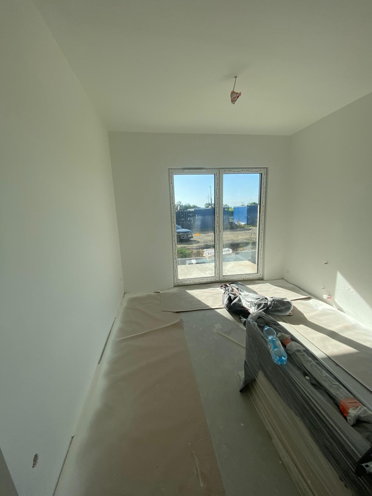 Wnętrze pokoju po malowaniu ścian na biało, z zabezpieczoną podłogą i widocznymi materiałami budowlanymi. Duże okno balkonowe z widokiem na zewnątrz.