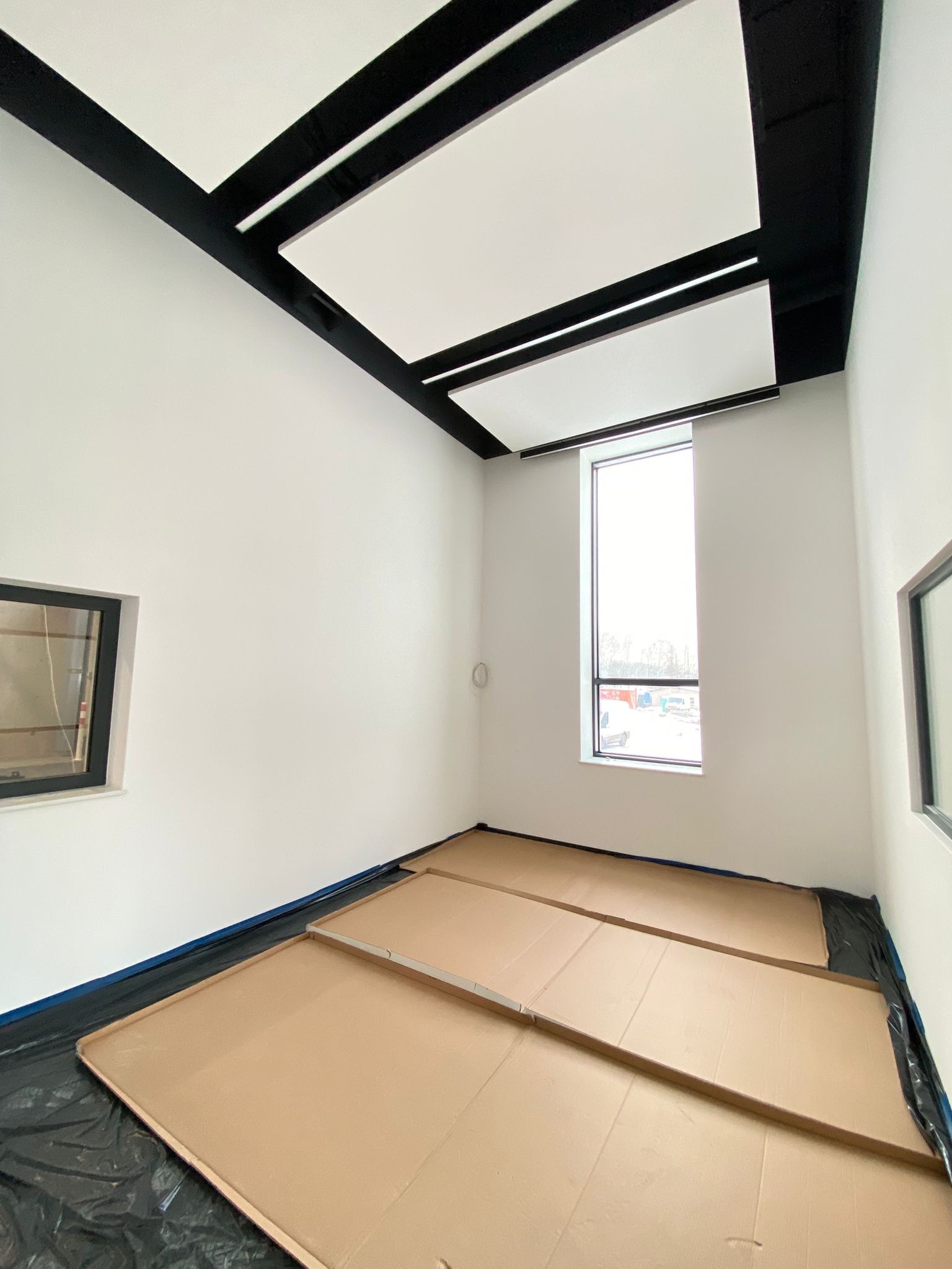 Pokój w trakcie przygotowań do malowania: zabezpieczona podłoga kartonem i folią, białe ściany, czarne elementy sufitu i ramy okienne. Widoczne okno i fragment drugiego okna.