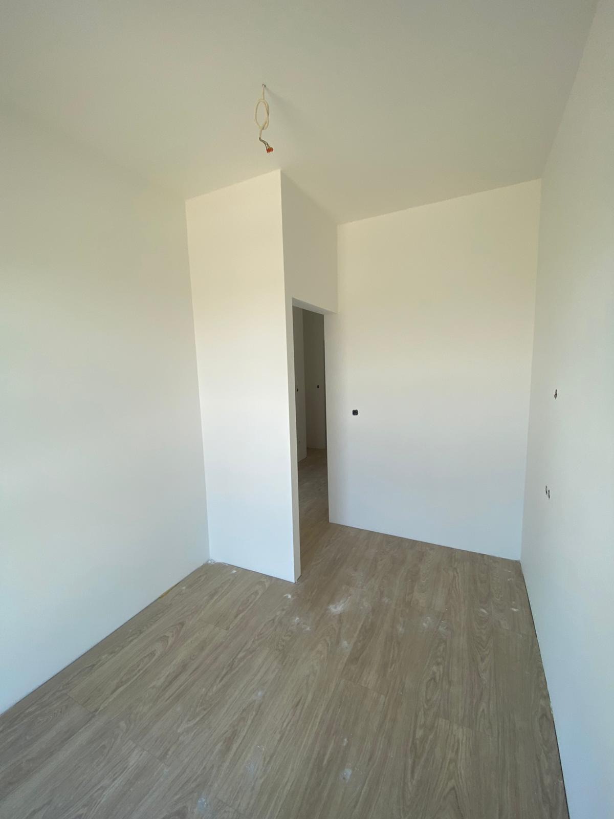 Pomalowane na biało wnętrze z panelami podłogowymi. Widoczne kable wystające z sufitu i fragment przejścia do innego pomieszczenia. Świeżo po malowaniu.