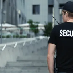 Mężczyzna w czarnej koszulce z napisem SECURITY na plecach, stojący tyłem na schodach przed nowoczesnym budynkiem, trzyma krótkofalówkę.