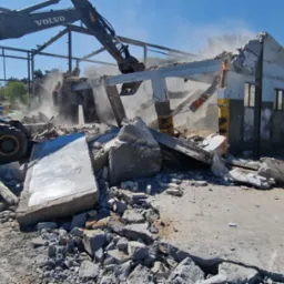 Żółta koparka Volvo demontuje fragment budynku z cegły i betonu, pozostawiając szkielet konstrukcji, w tle drzewa i błękitne niebo.