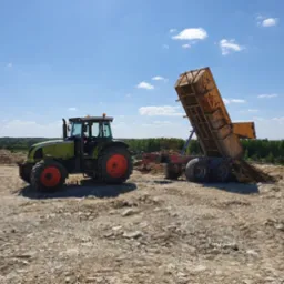 Zielony traktor Claas z pomarańczowymi felgami wyładowuje ziemię z przyczepy na placu budowy w słoneczny dzień.