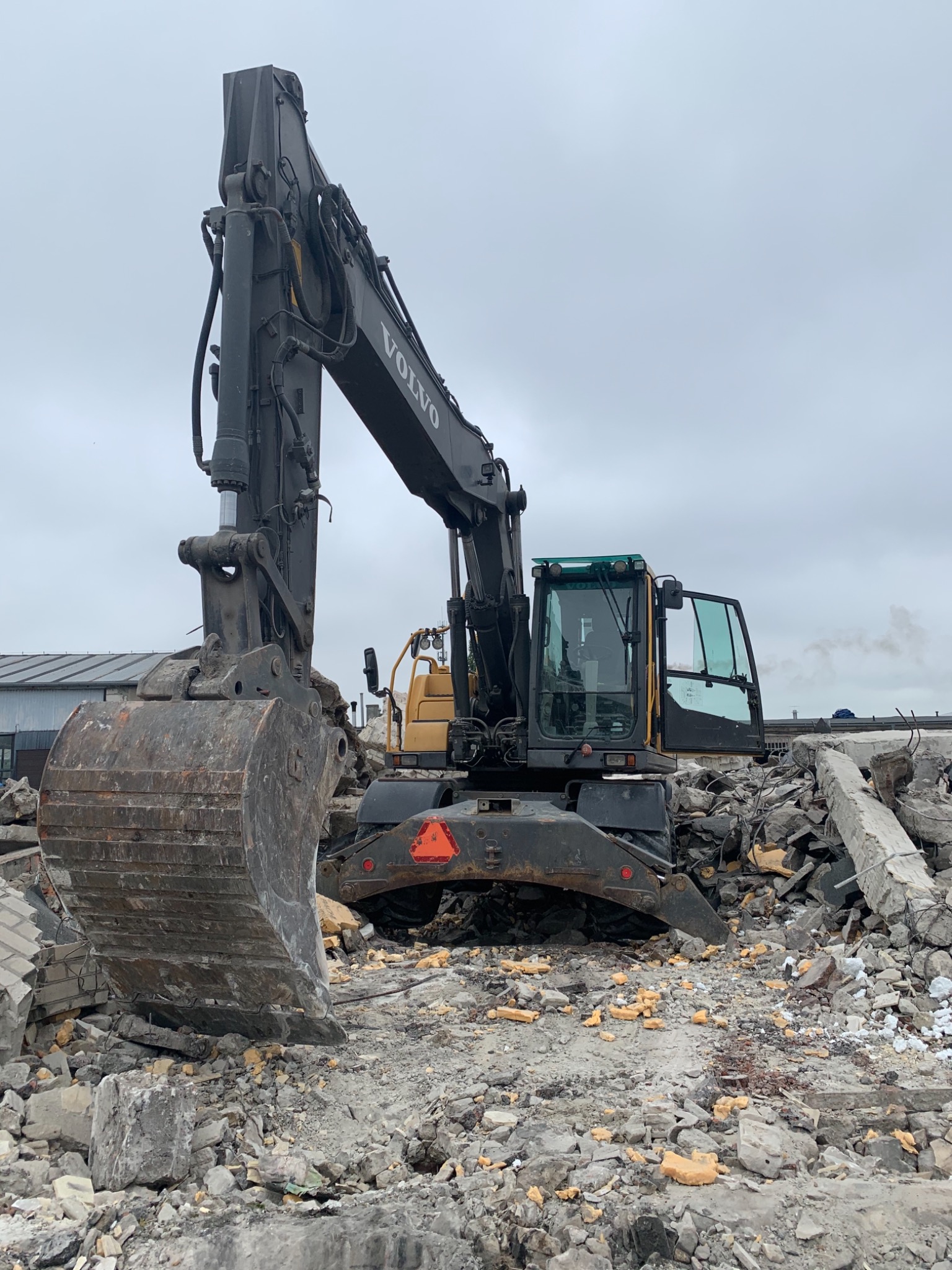 Czarny, używany, kołowy koparka Volvo z otwartymi drzwiami kabiny, stojąca na gruzowisku po rozbiórce budynku, z widocznym fragmentem innego pojazdu w tle.