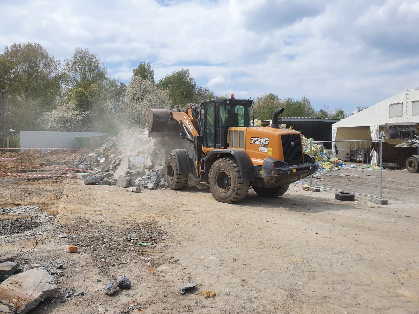 Pomarańczowa ładowarka CASE 721G nabiera gruz na placu budowy, w tle drzewa z białymi kwiatami i budynki gospodarcze.