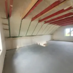 Poddasze po ociepleniu: biała pianka izolacyjna między czerwonymi belkami stropowymi, widoczne okno i surowa betonowa podłoga.